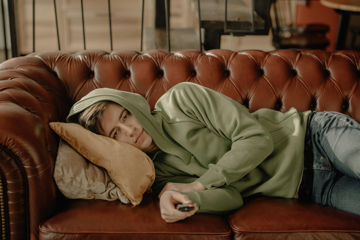 Bored young adult in hoodie lying on couch with game controller