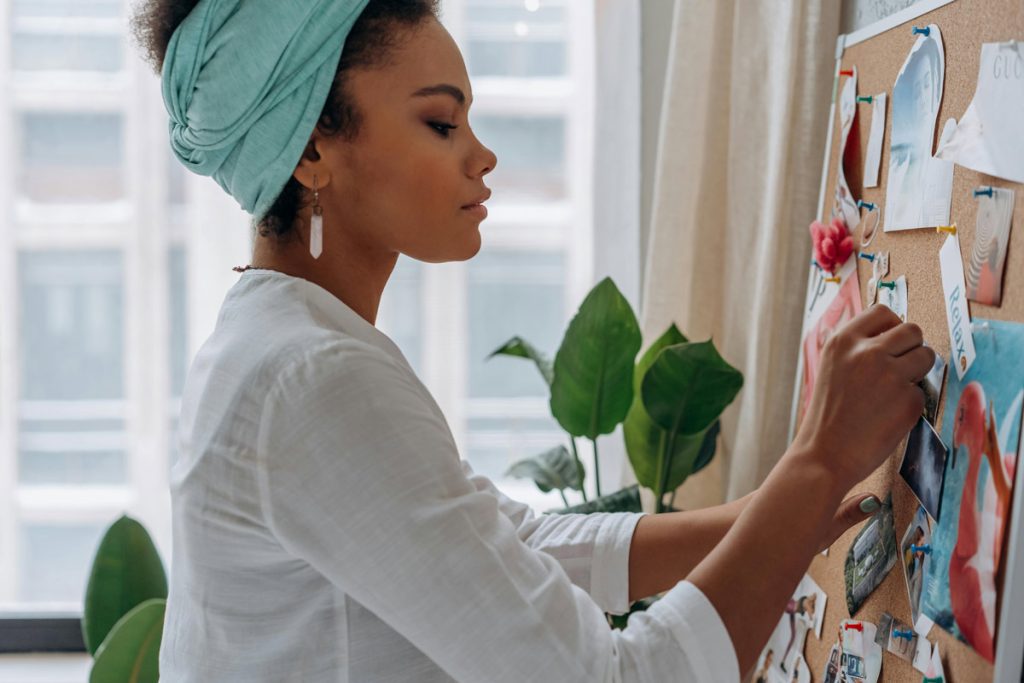 A woman in a light shirt and turquoise headscarf attaches photos to a corkboard.