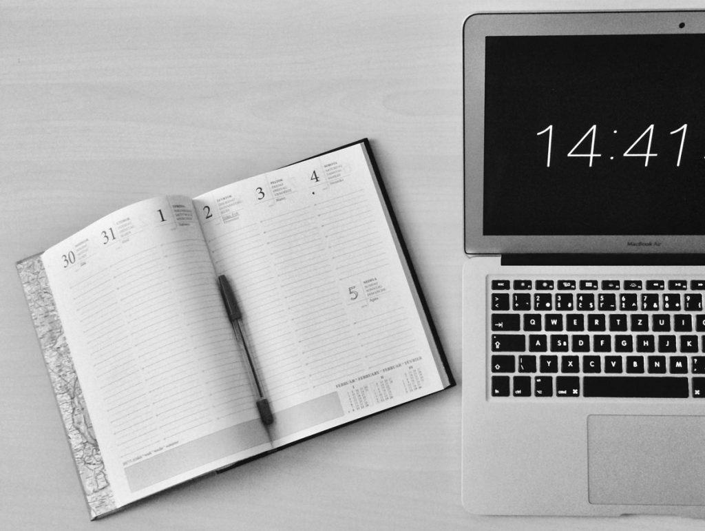  notebook with a pen on a table, showing dates and a laptop displaying the time.