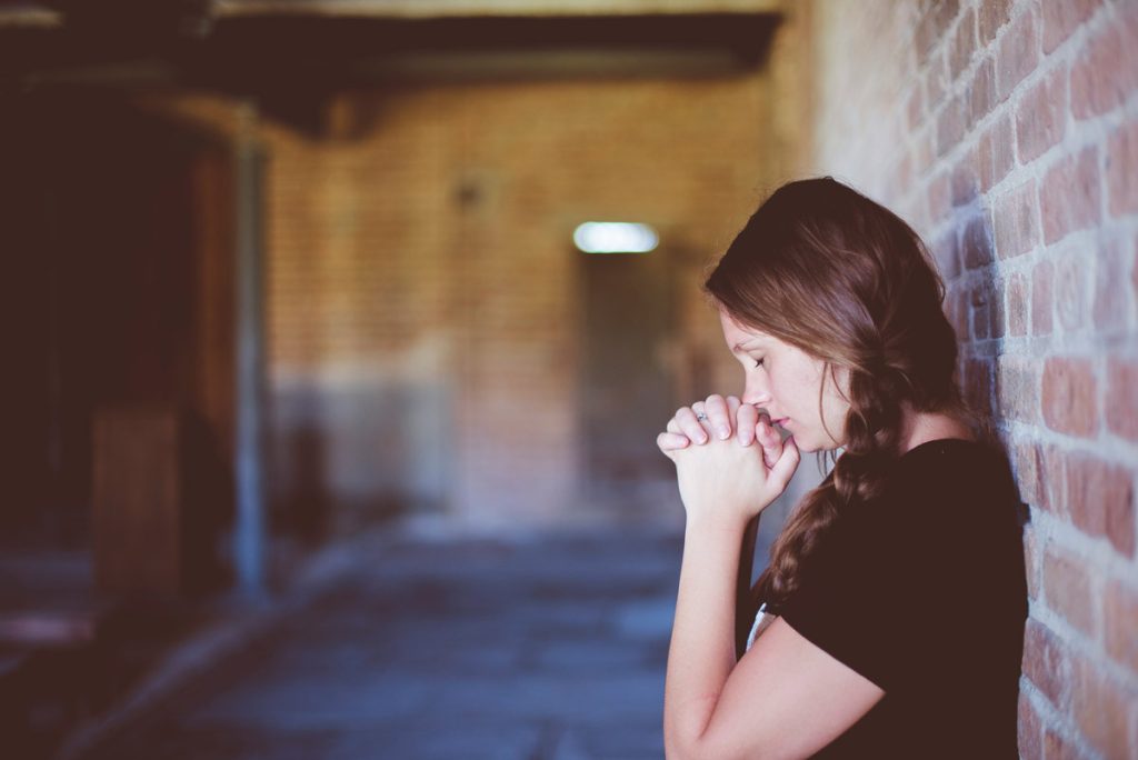 Young woman standing against wall, praying.